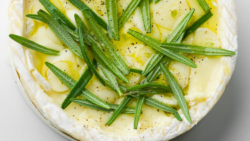 Wheel of Camembert pasta with rosemary leaves and sliced garlic.