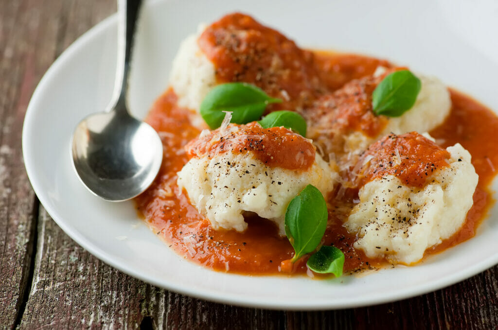 A plate of ricotta dumplings with tomato sauce and a spoon on the side.