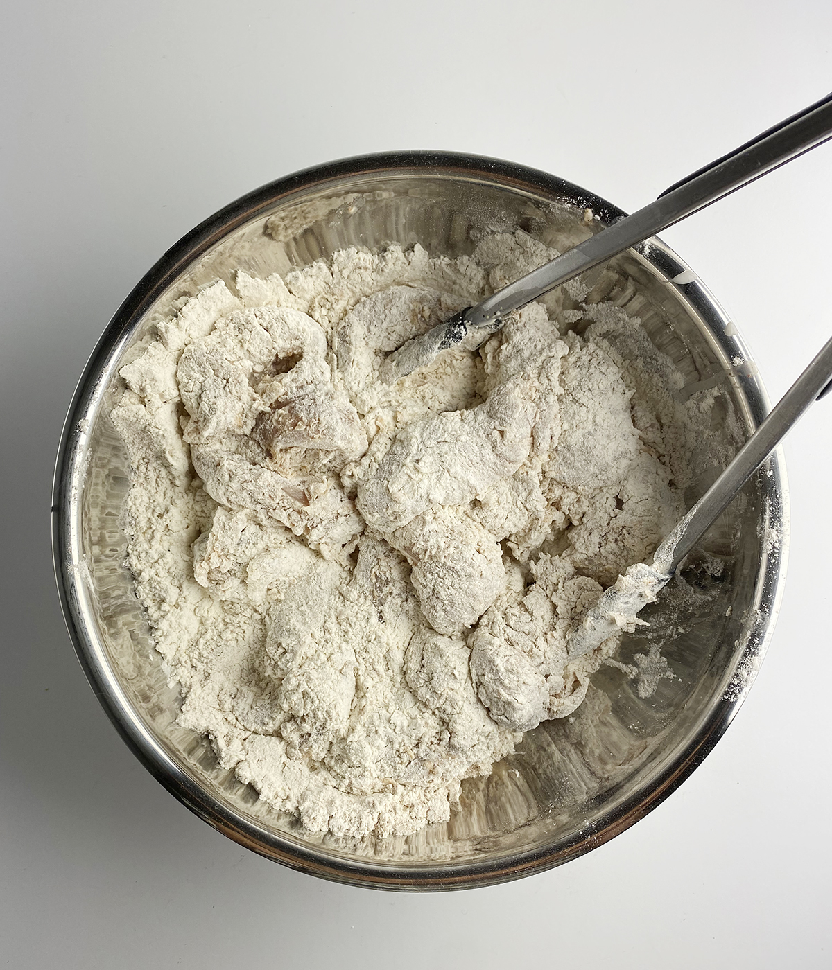 Chicken tenders being tossed with flour in a metal bowl with tongs.