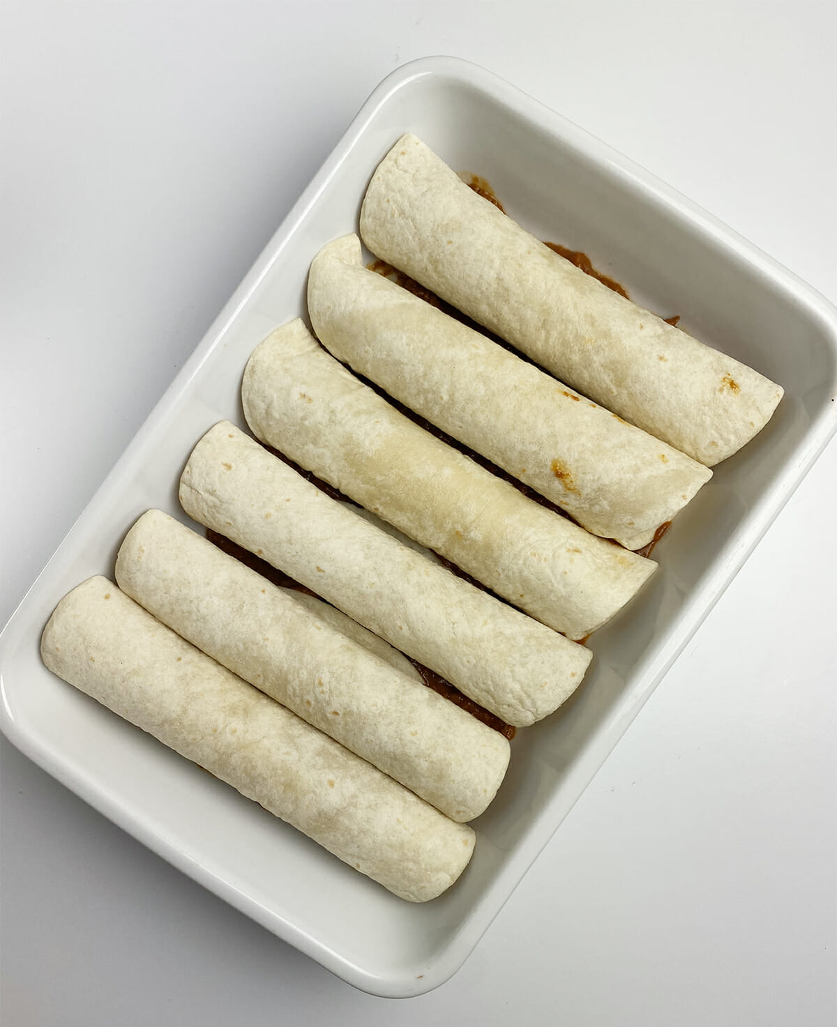 Hot dogs rolled up in flour tortillas on top of a layer of chili in a casserole dish.