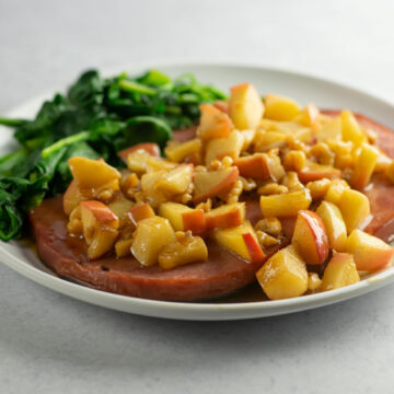 Ham steak with apples and walnut sauce on a plate with spinach on the side.