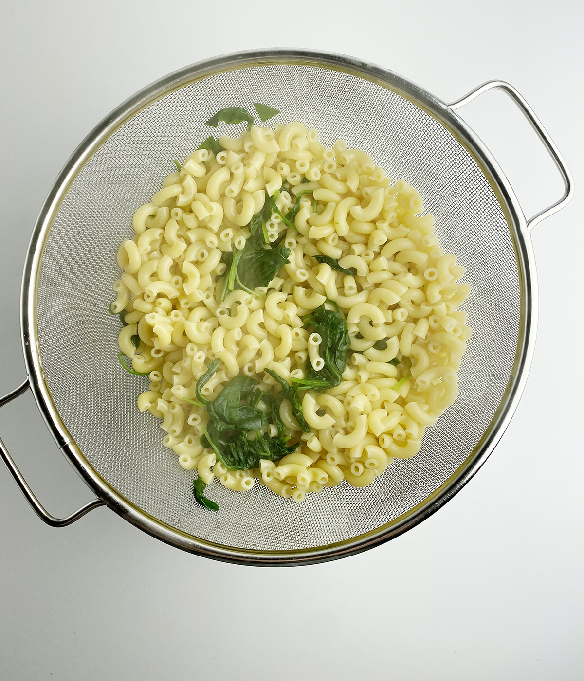 Cooked elbow macaroni and spinach draining in a strainer.